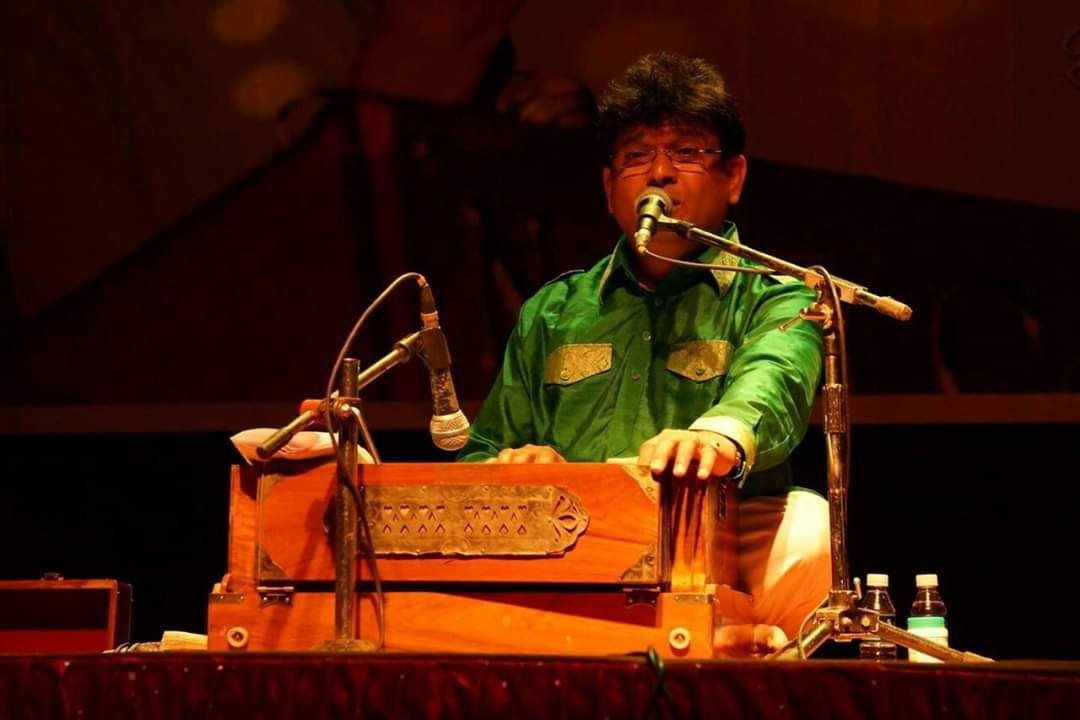Sanjay Banubakode singing at the harmonium under stage lights, green shirt