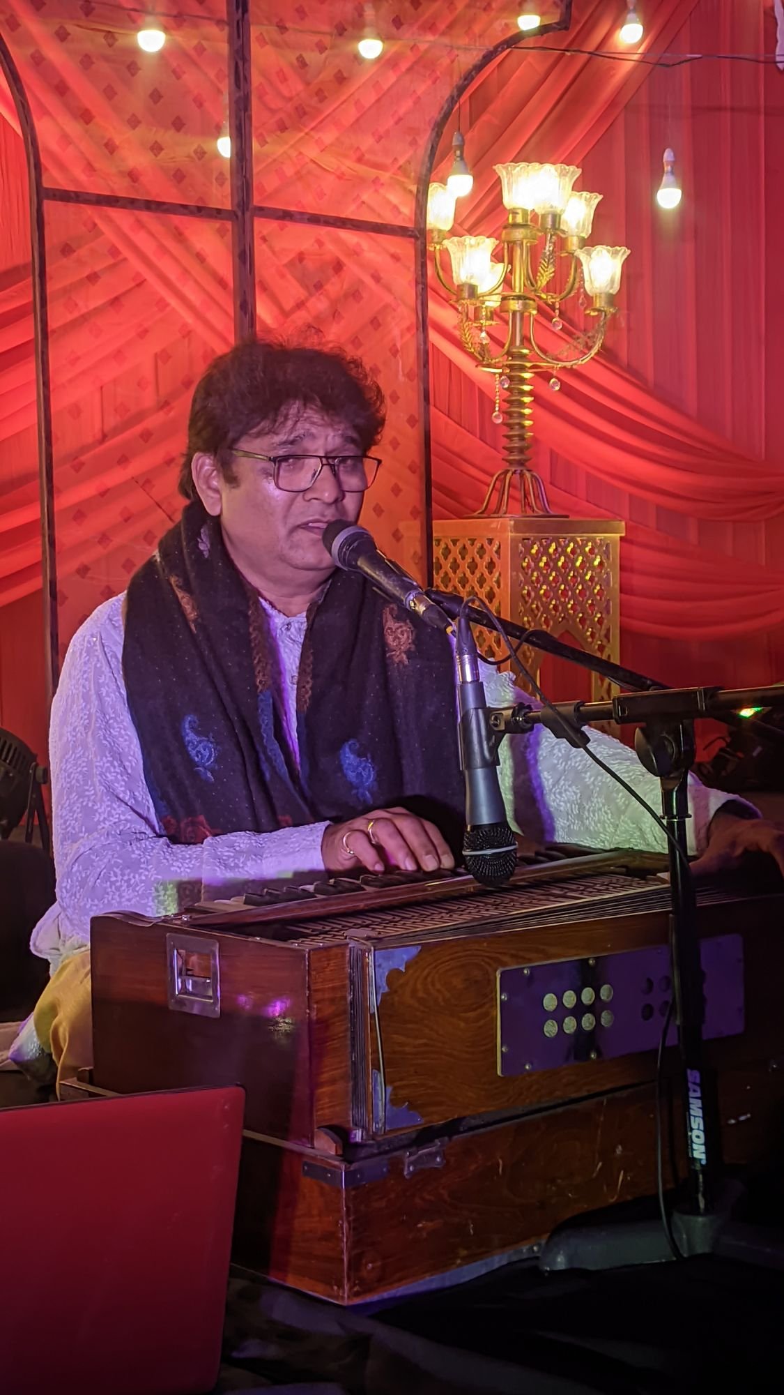 Sanjay Banubakode at the harmonium under a chandelier, red drapes behind