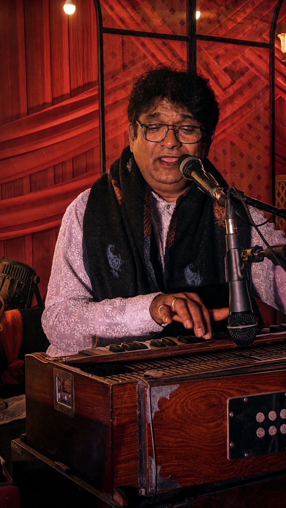Sanjay Banubakode singing at the harmonium in front of red drapes, black stole over a grey kurta