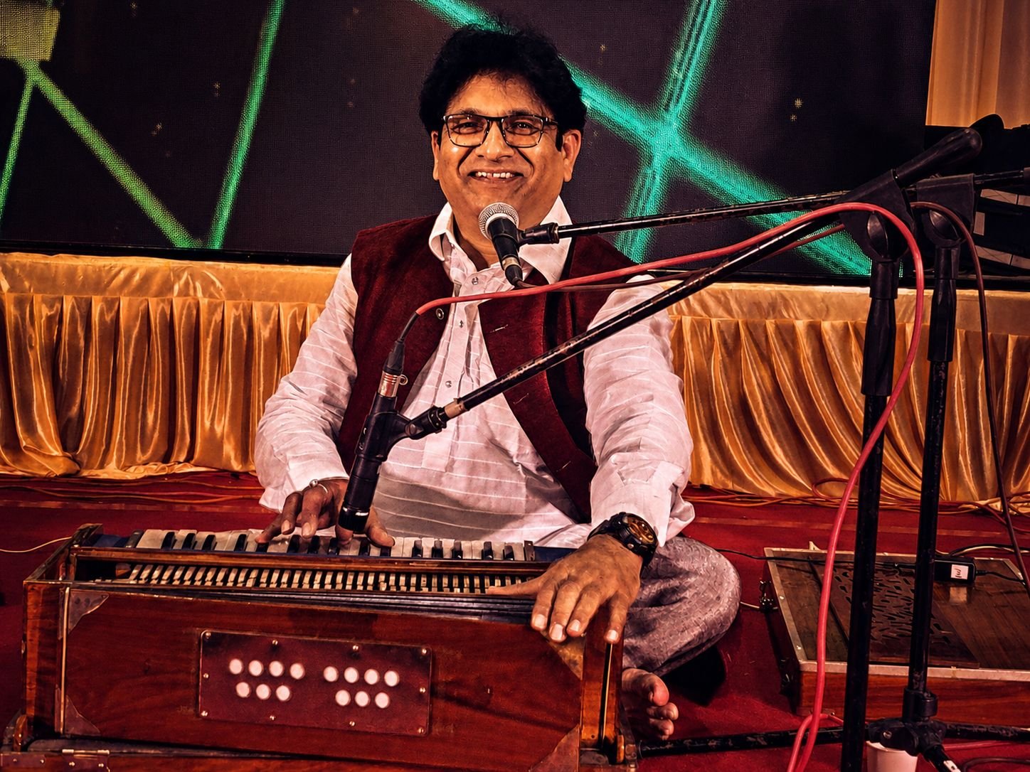 Sanjay Banubakode smiling at the harmonium, maroon jacket over white kurta, red curtain behind
