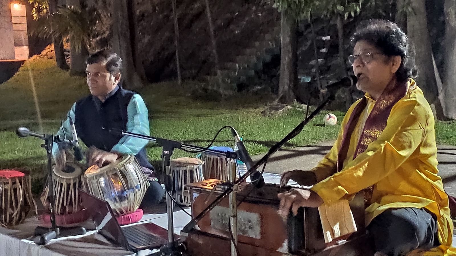 Sanjay Banubakode in yellow at the harmonium outdoors, tabla player beside him